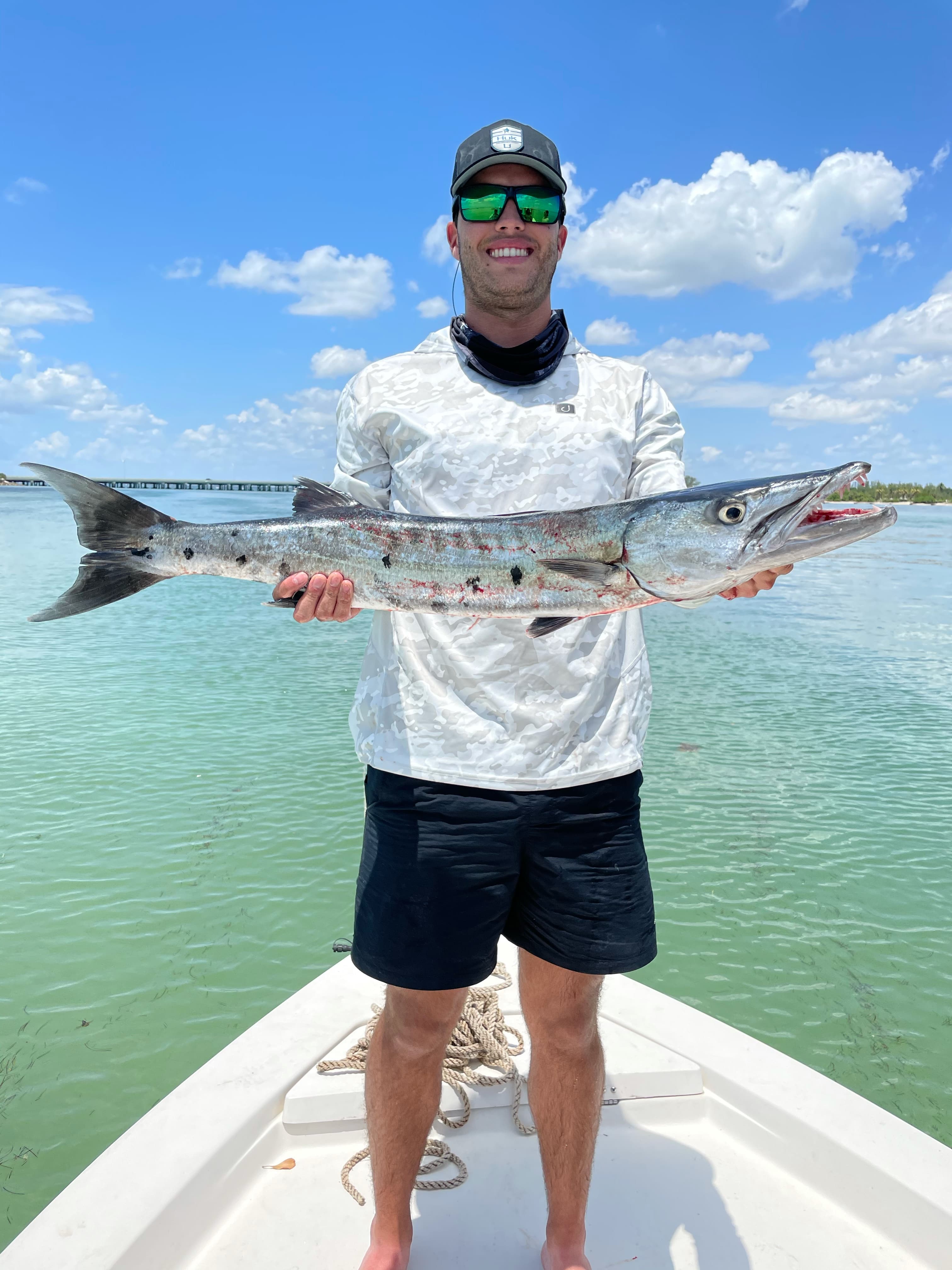 Angler on the bow holding a long barracuda over shallow turquoise water