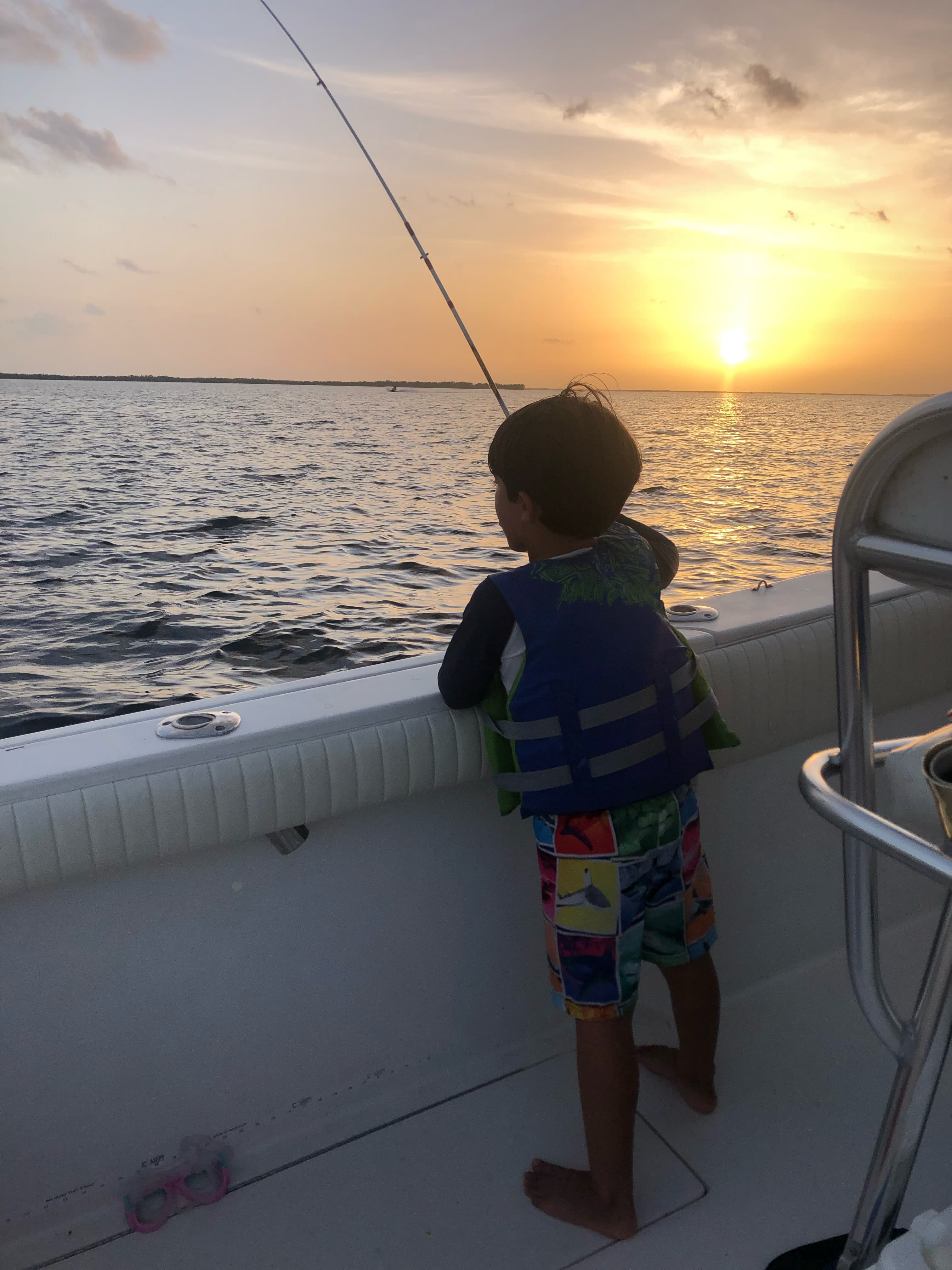 A child in a life jacket watching a fishing rod at sunset from the boat